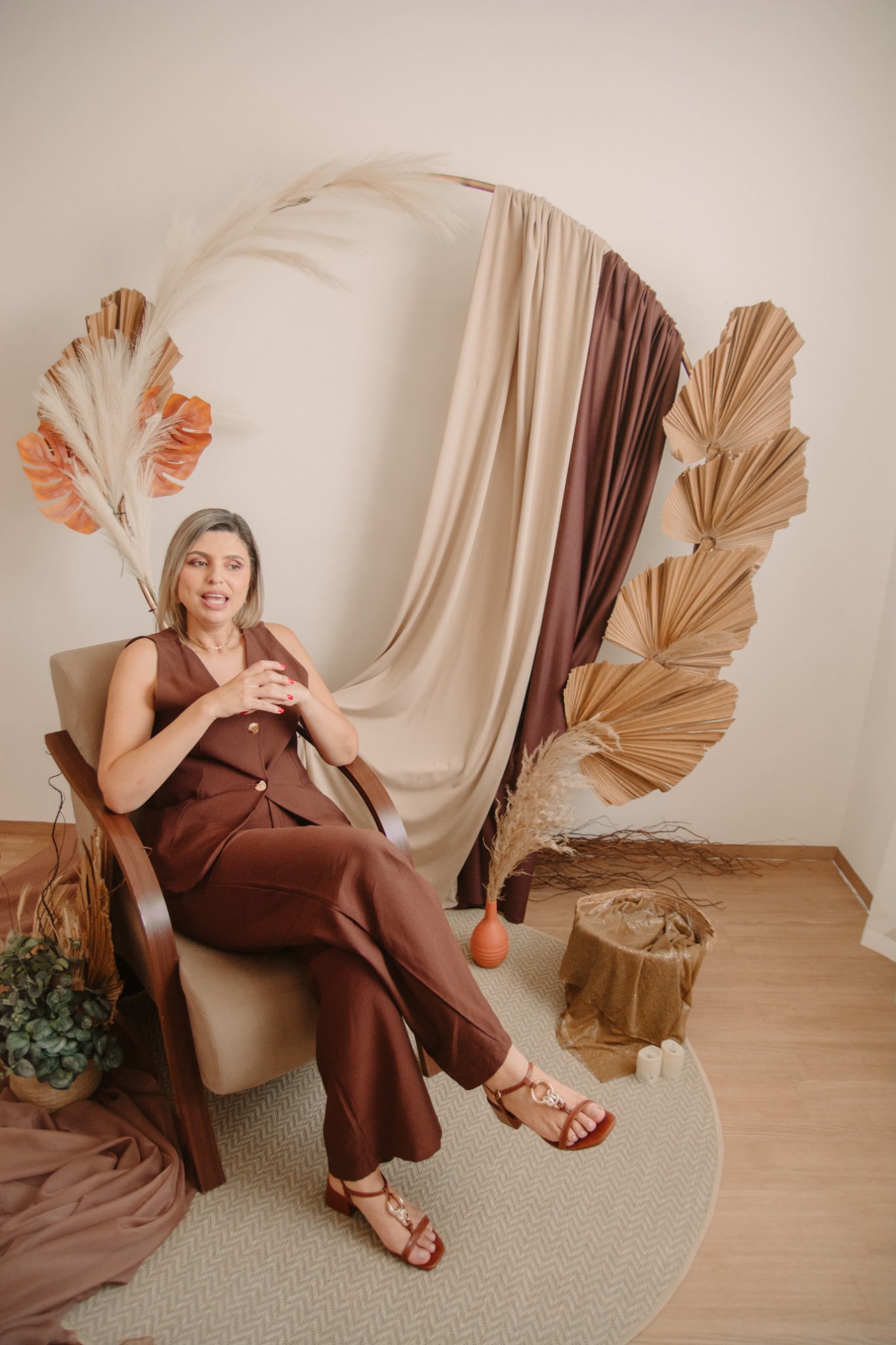 Thaís Guimarães sentada em poltrona, em cenário editorial de tons marrons e palhas secas