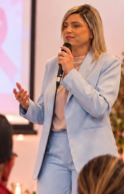 Thaís Guimarães de terno azul ao microfone em um ambiente decorado para Outubro Rosa