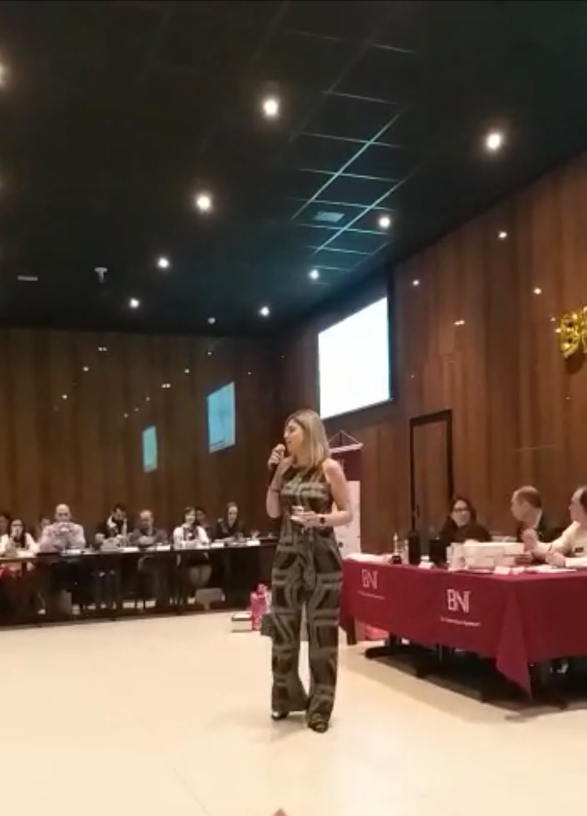 Thaís Guimarães palestrando no palco de um encontro do BNI, com a plateia em volta das mesas